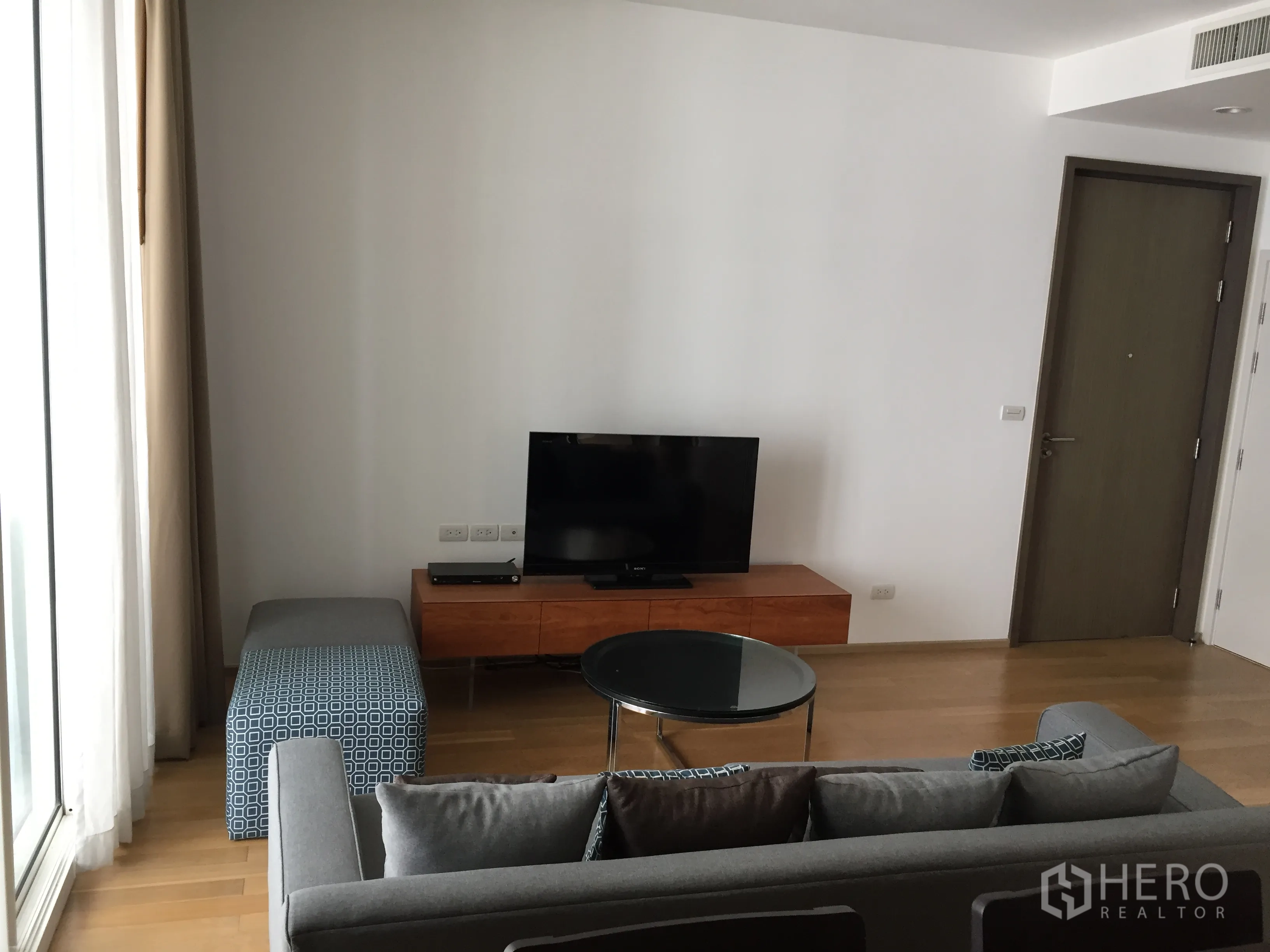 公寓 出租或出售在Watthana, Bangkok - 明亮客厅，灰色沙发、玻璃茶几、木质电视柜与落地窗帘。