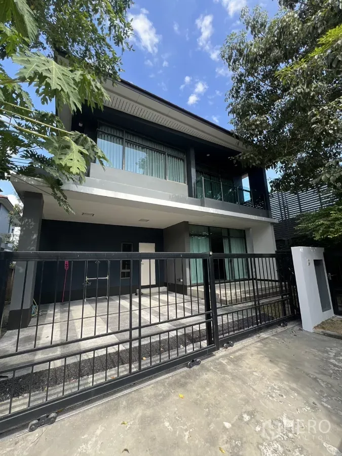 บ้าน ให้เช่าในBang Kapi, Bangkok - ด้านหน้าบ้านเดี่ยวสองชั้นสไตล์โมเดิร์น พร้อมรั้วเลื่อน ระเบียง และทางเข้ารถร่มรื่นด้วยต้นไม้