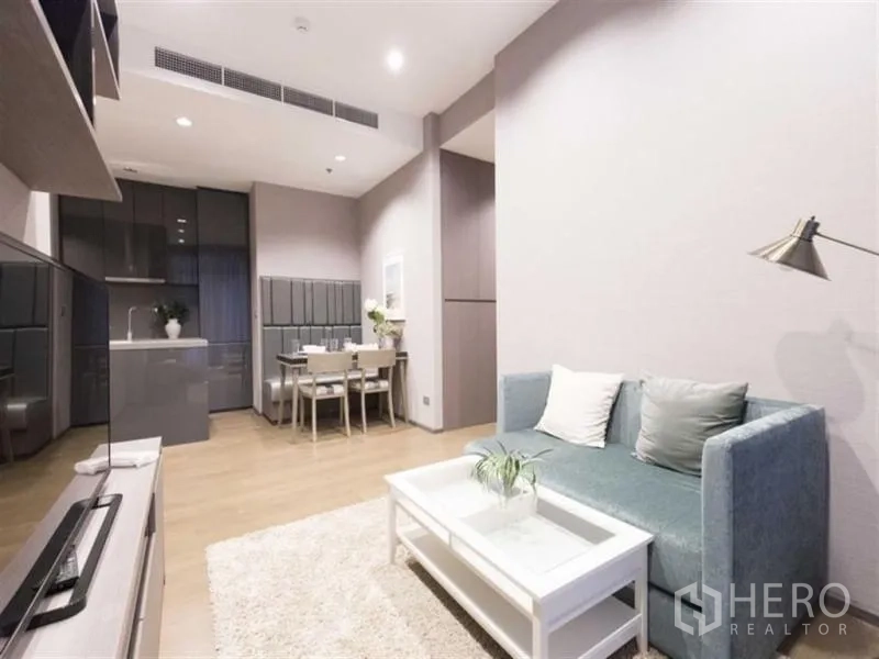 Condominium for rent in Bang Rak, Bangkok - Bright open-plan living room with teal sofa, white coffee table, dining set and sleek grey kitchen cabinetry on light wood floors.
