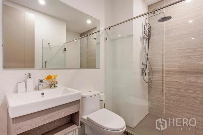 Condominium for rent in Watthana, Bangkok - Modern bathroom with glass-enclosed rain shower, vanity and toilet in light wood and white finishes.