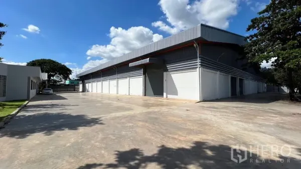 Warehouse for rent in Thanyaburi, Pathum Thani - Modern warehouse exterior in Thanyaburi with multiple roller shutters and a wide concrete yard under blue skies.
