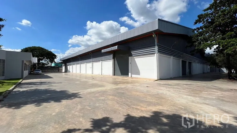 Warehouse for rent in Thanyaburi, Pathum Thani - Modern warehouse exterior in Thanyaburi with multiple roller shutters and a wide concrete yard under blue skies.