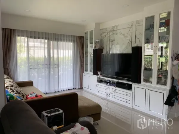Detached House for rent or sale in Samut Prakan - Bright living room with built-in TV wall, large windows and sheer curtains.