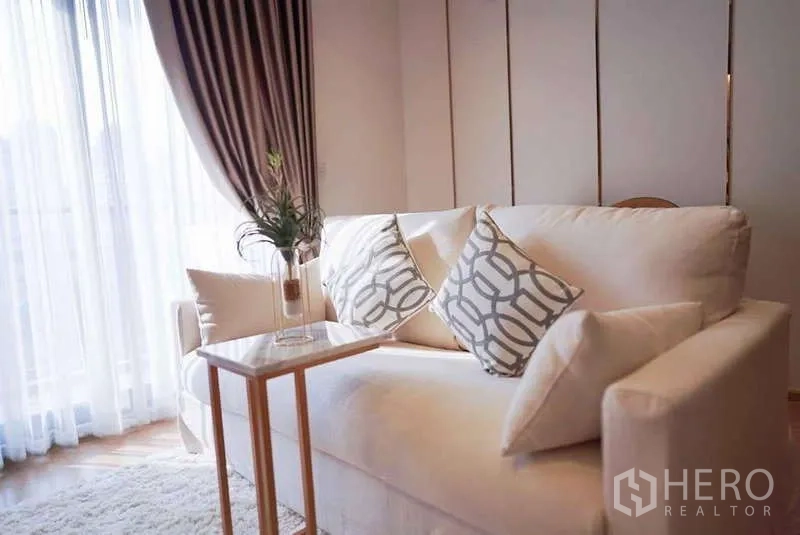 Condominium for rent or sale in Watthana, Bangkok - Living area by sheer balcony curtains featuring a cream sofa, geometric cushions, and slim side table.