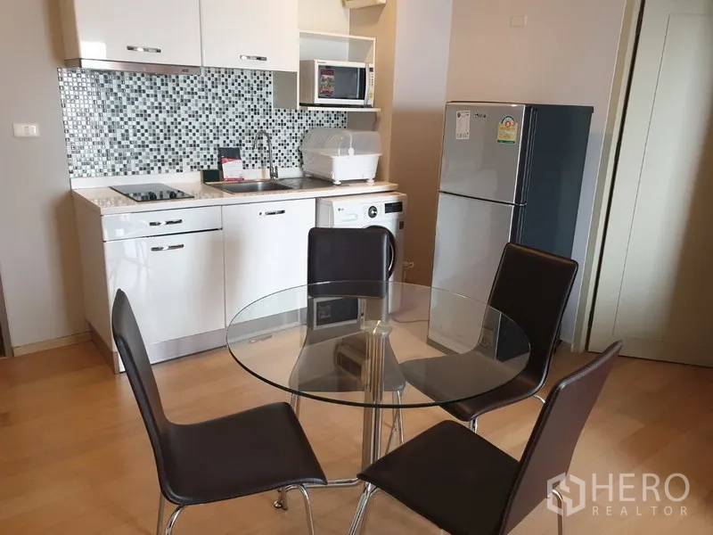 คอนโด ให้เช่าในWatthana, Bangkok - ครัวสีขาวกะทัดรัด ผนังกระเบื้องโมเสก เตาไฟฟ้า ตู้เย็น และโต๊ะกินข้าวกระจกทรงกลม