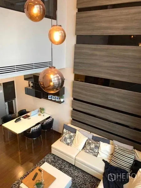 Condominium for rent or sale in Huai Khwang, Bangkok - Double-height living room with floor-to-ceiling windows, pendant lights, and a sectional sofa beside the dining area.