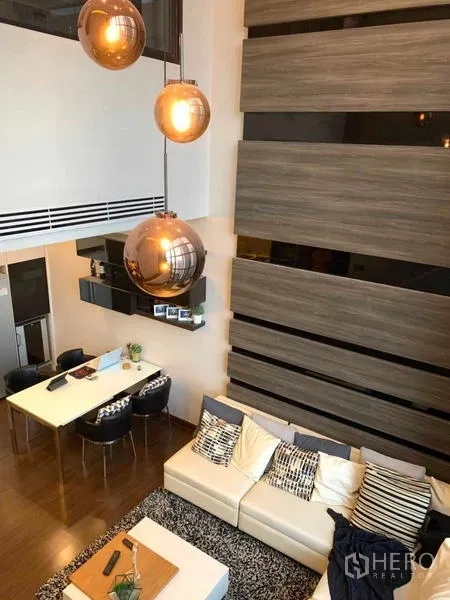 Condominium for rent or sale in Huai Khwang, Bangkok - Double-height living room with floor-to-ceiling windows, pendant lights, and a sectional sofa beside the dining area.