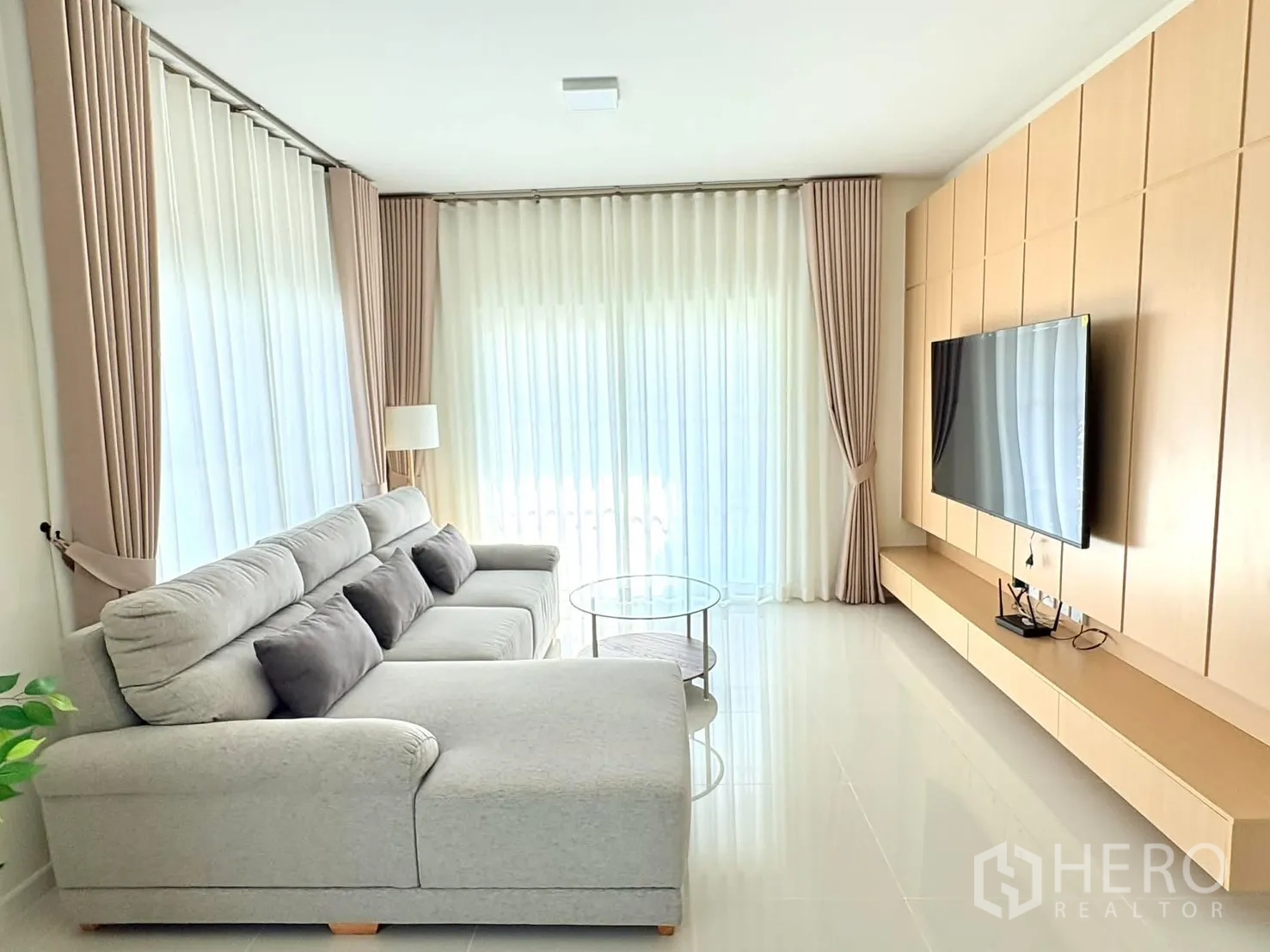 独立式住宅 for rent in Bang Phli, Samut Prakan - 客厅视角可见大型电视墙、转角沙发和整面窗帘。