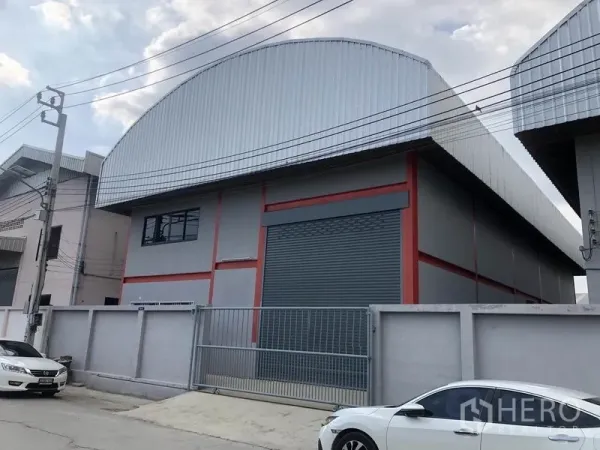 货栈 出租在Nonthaburi - 街景视角的仓库，带围栏与大门，大幅卷闸门与金属外墙，院内可停车。