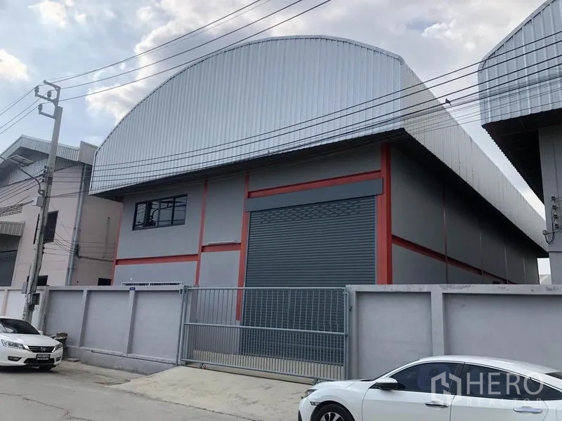 货栈 出租在Nonthaburi - 街景视角的仓库，带围栏与大门，大幅卷闸门与金属外墙，院内可停车。