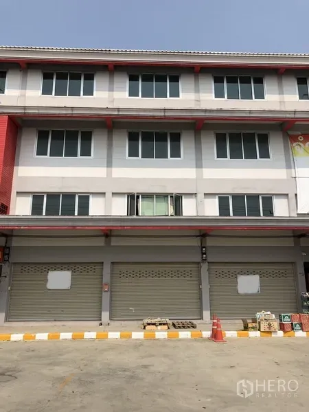 อาคารพาณิชย์ ให้เช่าในLam Luk Ka, Pathum Thani - อาคารพาณิชย์หลายชั้น ผนังสีเทา หน้าต่างบานใหญ่ ประตูม้วนโลหะ และพื้นที่จอดรถด้านหน้า