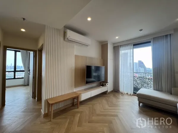 Condominium for rent or sale in Chatuchak, Bangkok - Bright living room showing doorway to bedroom and balcony with sheer curtains and a built‑in TV wall.