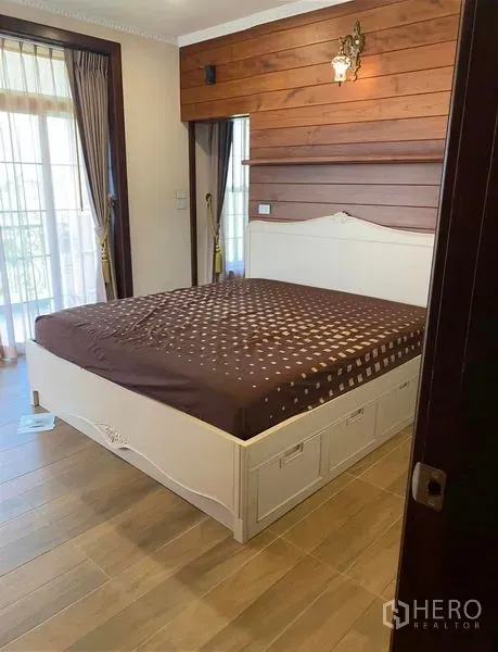 Detached House for sale in Bang Bo, Samut Prakan - Cozy bedroom with a white bed frame, wood accent wall, and balcony doors with curtains.
