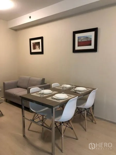 Condominium for rent or sale in Huai Khwang, Bangkok - Four-seat dining table beside a gray sofa with framed artwork on beige walls under soft lighting.