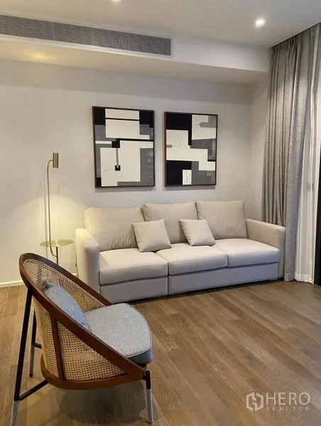 Condominium for rent in Pathum Wan, Bangkok - Sitting area with cream sofa, abstract wall art, accent chair, floor lamp, and full-length curtains.