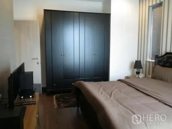 Condominium for rent or sale in Huai Khwang, Bangkok - Master bedroom with upholstered bed, large dark wardrobe, bedside lamp and window.