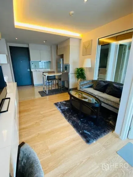 คอนโด ให้เช่าในWatthana, Bangkok - ห้องนั่งเล่นเปิดโล่ง พื้นไม้ ผนังกระจกกั้นห้องนอน พรม โซฟา และครัวขนาดกะทัดรัดพร้อมเคาน์เตอร์บาร์