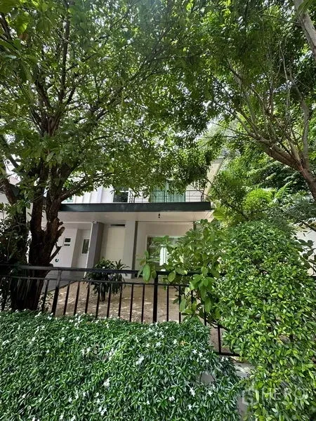 独立式住宅 出售在Prawet, Bangkok - 被茂密树荫环绕的房屋立面，带阳台与花园围栏（曼谷帕拉威）。
