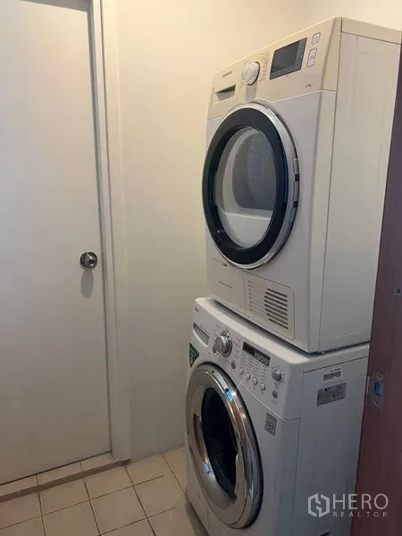คอนโด ให้เช่าในBangkok - มุมซักรีดพร้อมเครื่องซักผ้าและเครื่องอบผ้าตั้งซ้อน พื้นกระเบื้องและผนังสีขาว