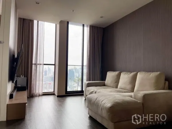 Condominium for rent in Pathum Wan, Bangkok - Cozy lounge with L-shaped sofa, TV unit, and tall windows showing a city skyline view.
