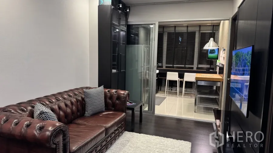 公寓 出租或出售在Khlong Toei, Bangkok - 客厅配棕色切斯特菲尔德皮沙发、地毯，电视墙与玻璃移门通向厨房。