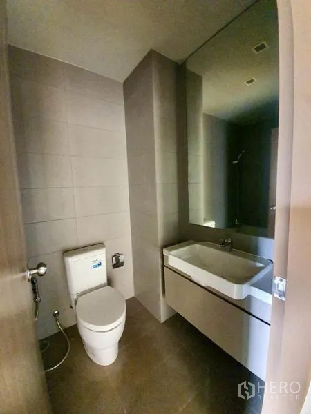 Condominium for sale in Ratchathewi, Bangkok - Modern bathroom with wall-mounted sink, large mirror and toilet in neutral tiles.