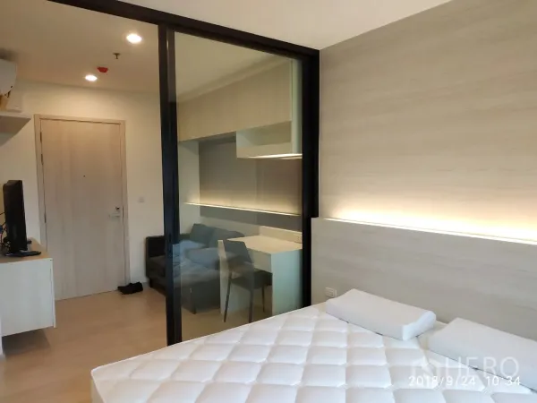 Condominium for rent in Huai Khwang, Bangkok - Bedroom view to living area through sliding glass wall with study table and sofa in light wood tones.
