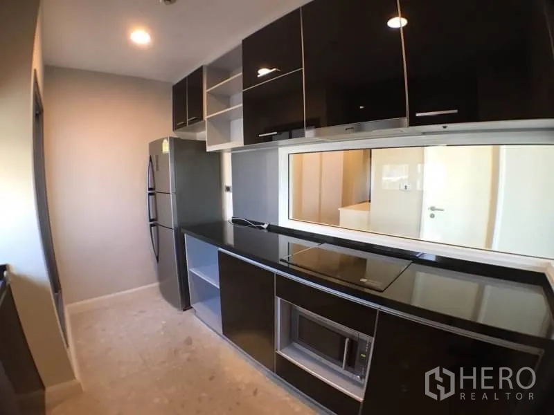 Condominium for rent in Khlong Toei, Bangkok - Enclosed galley kitchen with glossy black cabinets, cooktop and full-size refrigerator.