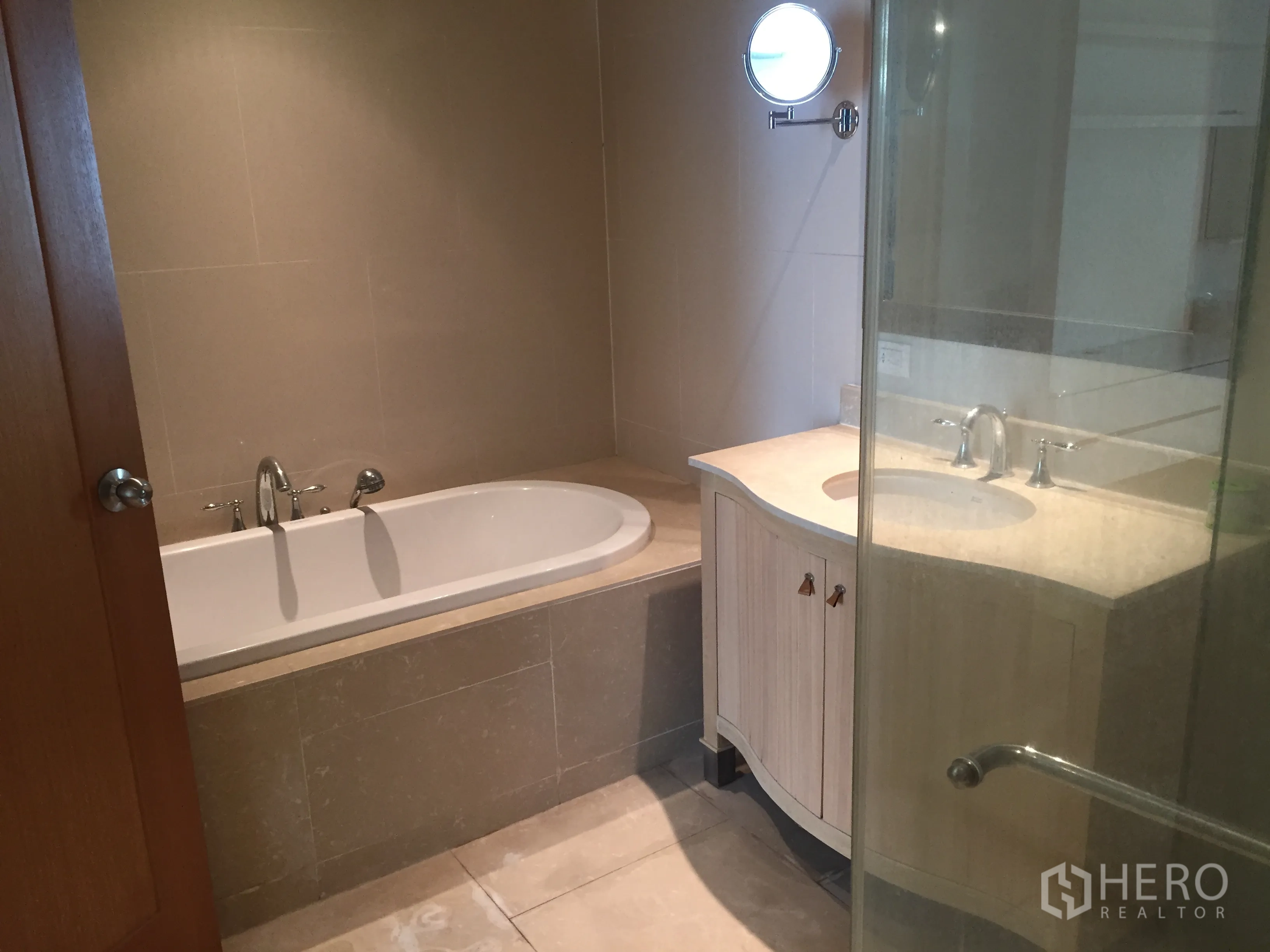 Condominium for rent in Sathon, Bangkok - Modern bathroom featuring a built-in bathtub, glass shower enclosure, and curved vanity in neutral tiles.