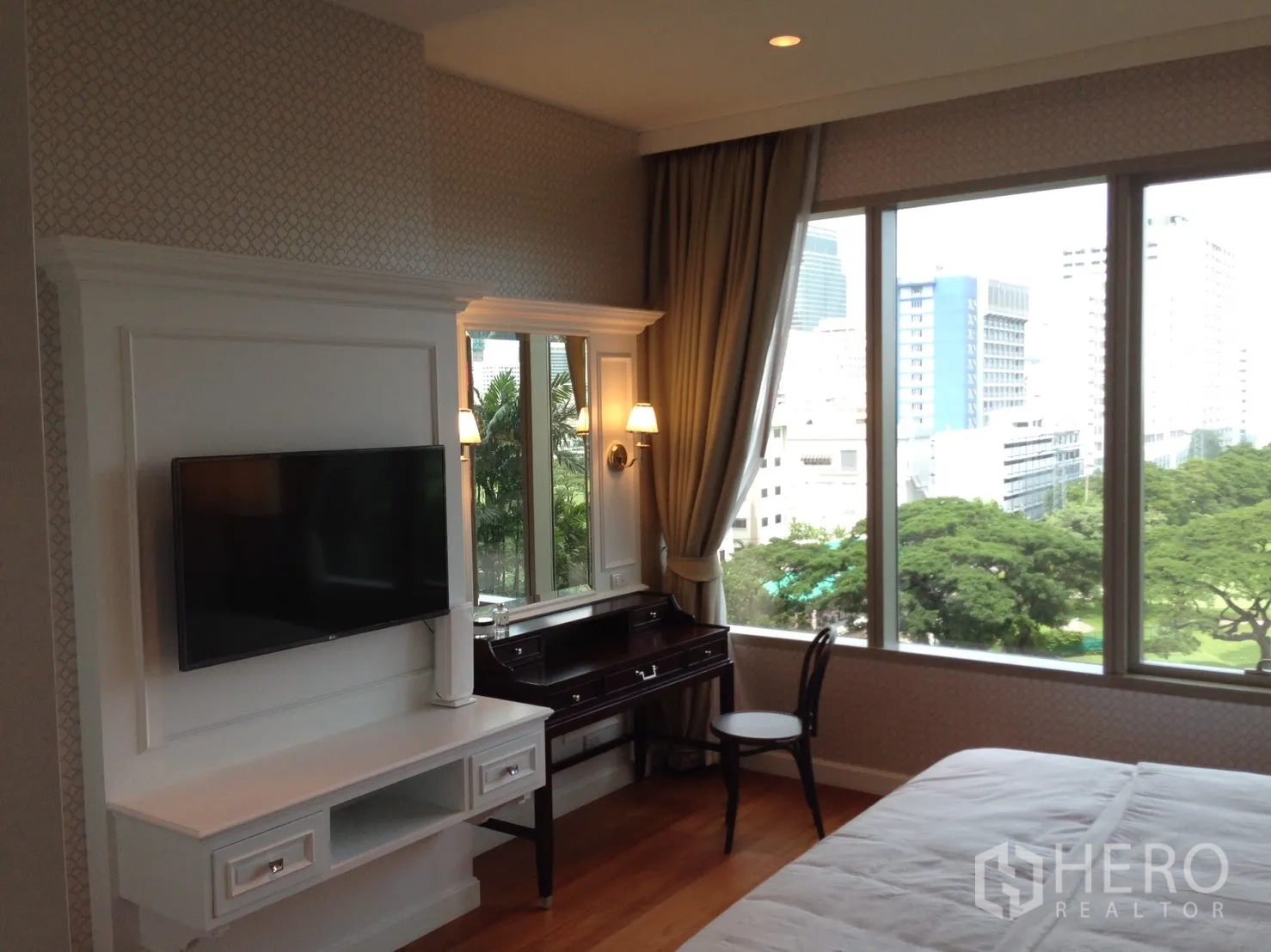 คอนโด ให้เช่าหรือขายในPathum Wan, Bangkok - มุมห้องนอนทีวีติดผนัง โต๊ะทำงาน และหน้าต่างสูงจากพื้นจรดฝ้า มองเห็นต้นไม้.