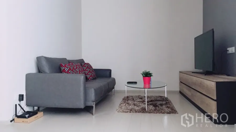 Condominium for rent in Pathum Wan, Bangkok - Minimalist living room with a grey sofa, patterned cushions, glass coffee table, rug, and TV console.