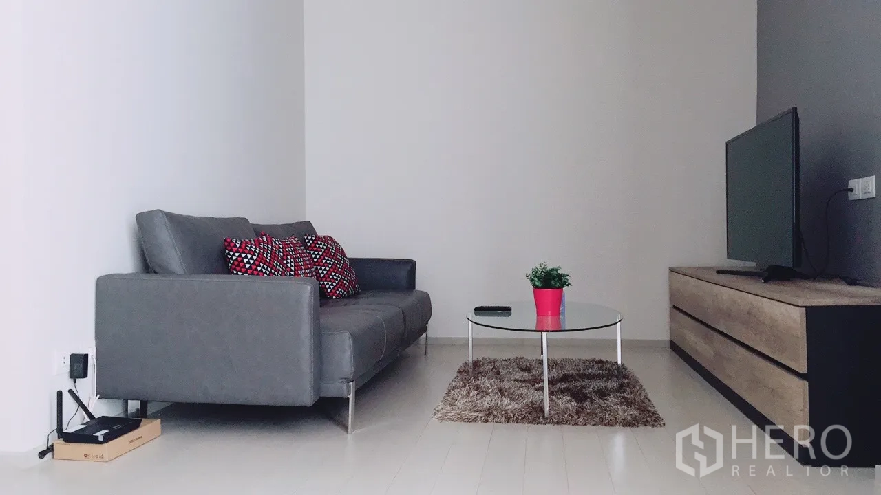 Condominium for rent in Pathum Wan, Bangkok - Minimalist living room with a grey sofa, patterned cushions, glass coffee table, rug, and TV console.