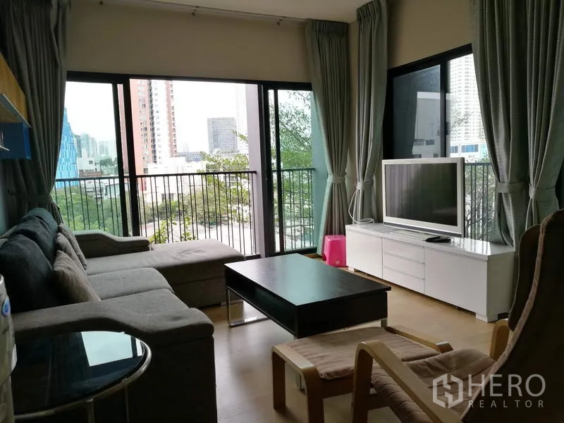 Condominium for rent in Watthana, Bangkok - Living room with L-shaped sofa, coffee table, TV on white console and wraparound windows opening to a balcony with city views.