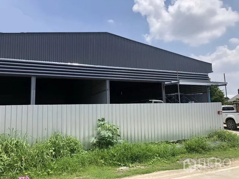Warehouse for rent in Bang Khen, Nonthaburi - Side view of a large grey metal warehouse behind a temporary fence and grass verge.