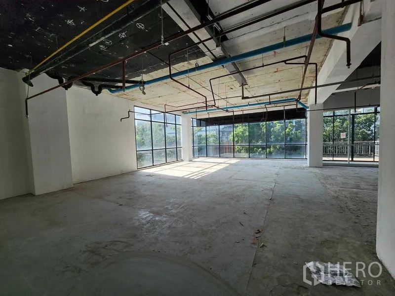 Retail Space for rent in Sathon, Bangkok - Sunlit shell retail corner with floor-to-ceiling windows, exposed pipes and concrete floor overlooking greenery.