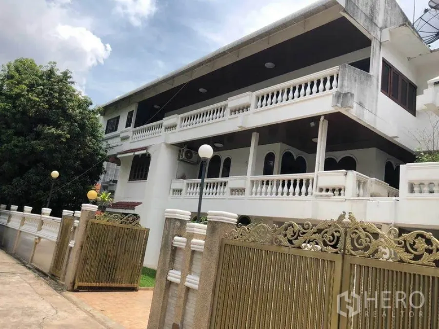 独立式住宅 出租在Watthana, Bangkok - 两层独栋别墅外观，白色扶栏与华丽金色大门，天空多云。