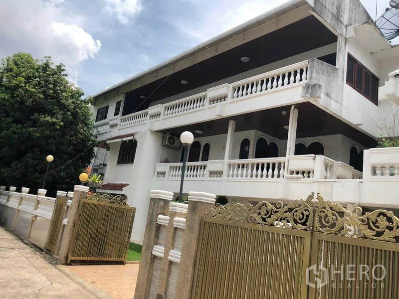 独立式住宅 出租在Watthana, Bangkok - 两层独栋别墅外观，白色扶栏与华丽金色大门，天空多云。