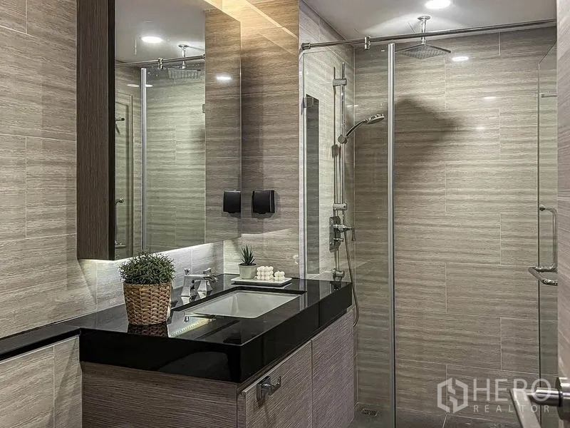 Condominium for rent in Pathum Wan, Bangkok - Contemporary bathroom with glass-enclosed shower, rain head, and black stone vanity.