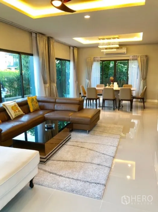 Detached House for rent in Bang Kapi, Bangkok - Open-plan living and dining area with L-shaped sofa, recessed lighting and greenery outside.
