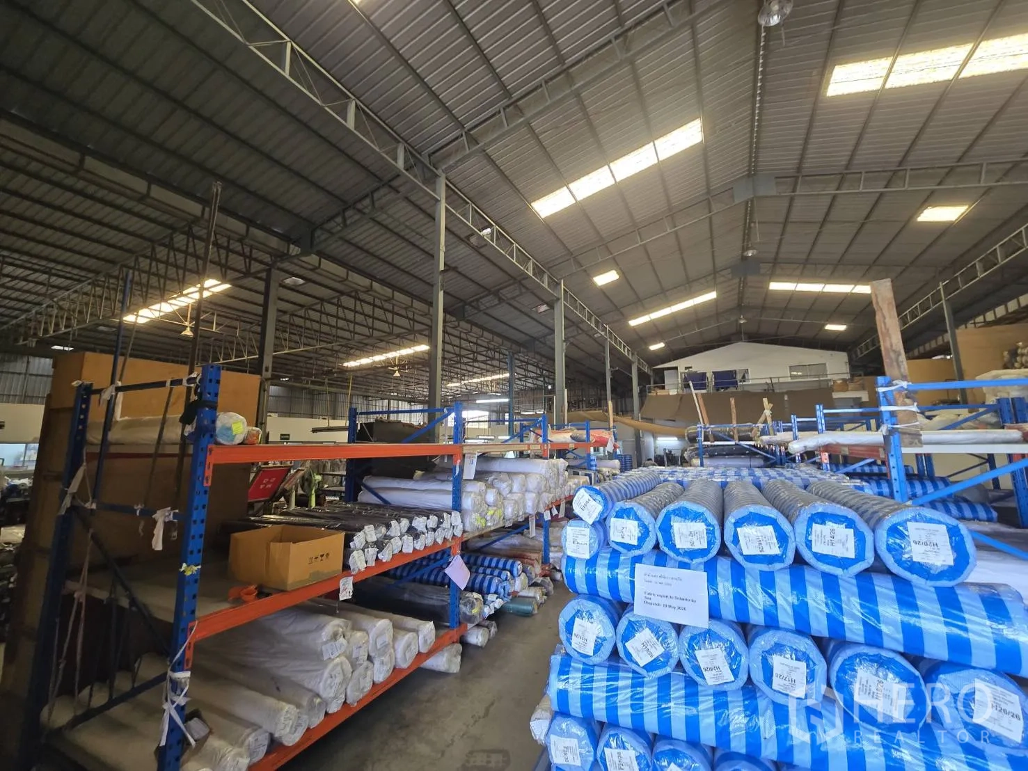 Factory for rent in Sam Phran, Nakhon Pathom - Spacious warehouse interior with high steel roof, racks of fabric rolls, and mezzanine office.
