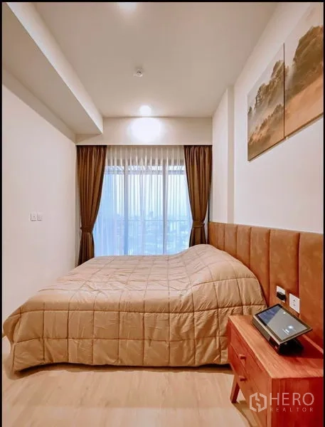 Condominium for sale in Huai Khwang, Bangkok - Cozy bedroom with padded headboard, bedside table and large window showing a city view.