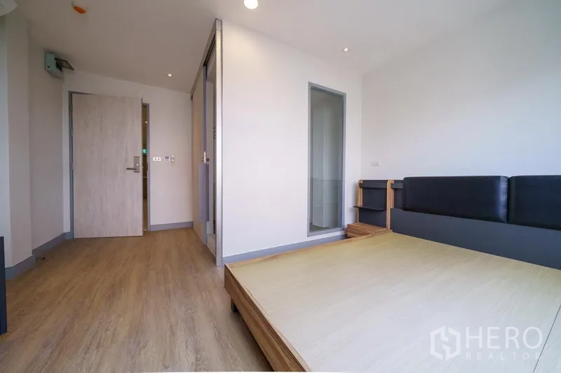 Home Office for rent in Khlong Toei, Bangkok - Compact bedroom with light wood floors, upholstered bed and a sliding glass door leading to an ensuite.