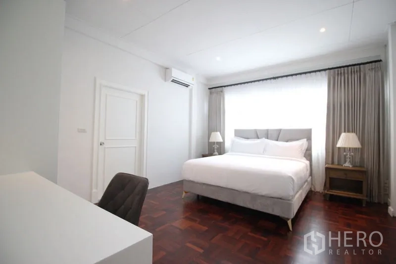 Detached House for rent in Chatuchak, Bangkok - Bright bedroom with parquet wood flooring, gray upholstered bed, desk, large curtained window, and air conditioning.