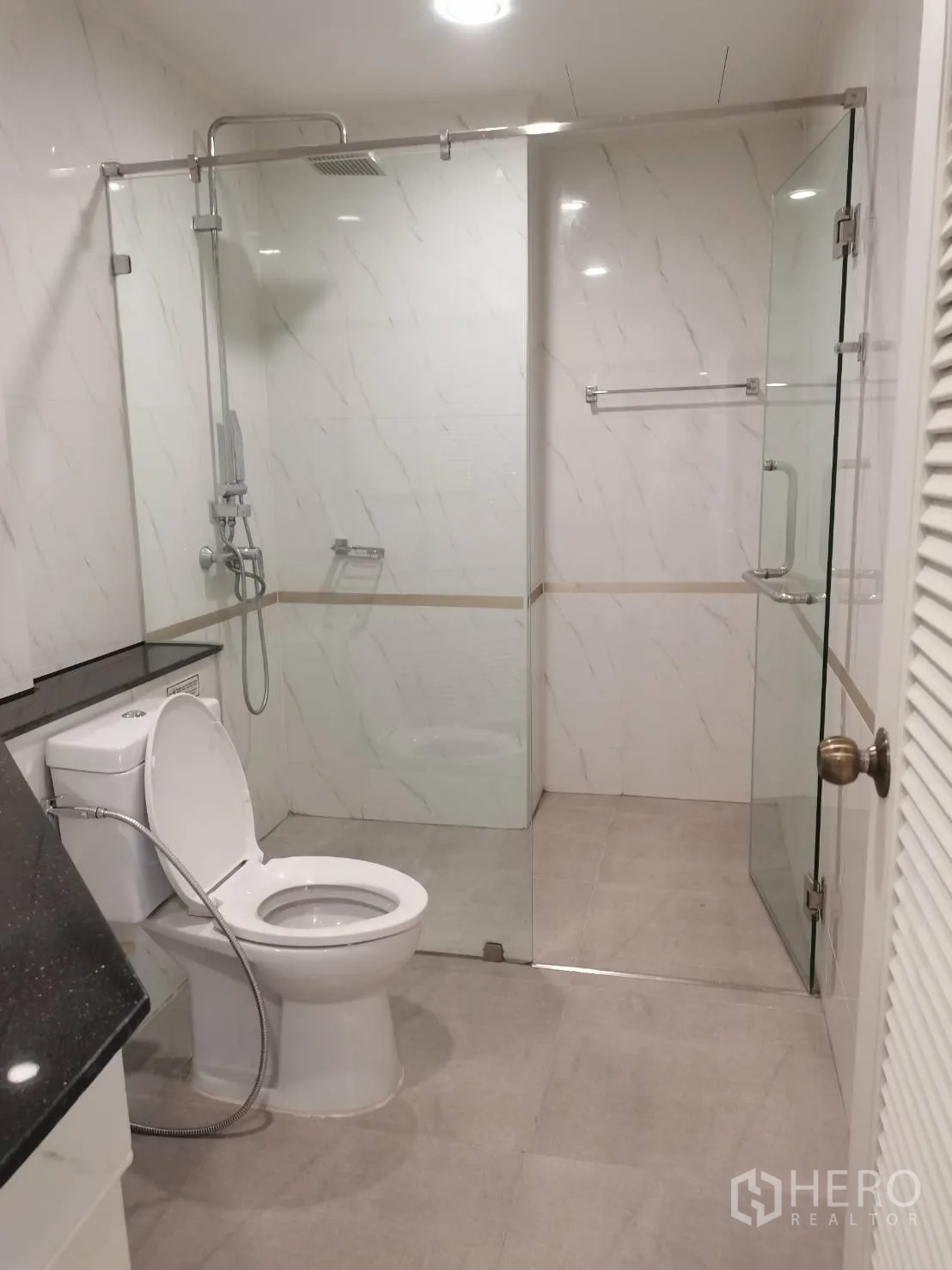 Condominium for rent in Watthana, Bangkok - Modern bathroom with glass-enclosed rain shower, toilet, and vanity with black countertop.