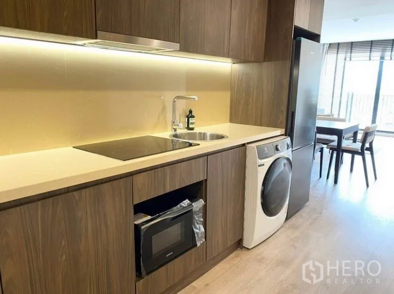 Condominium for rent in Pathum Wan, Bangkok - Modern galley kitchen with wood cabinets, induction cooktop, sink, microwave, washer and tall fridge.