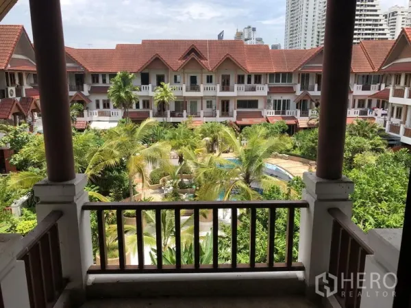 Townhouse for rent or sale in Watthana, Bangkok - Balcony overlooking a tropical courtyard garden with a shared swimming pool and red-roof townhouse compound.