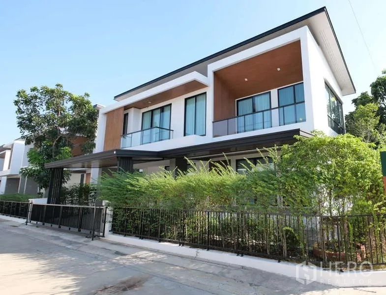 Detached House for rent or sale in Thanyaburi, Pathum Thani - Modern two-storey detached house exterior with glass balconies, corner greenery and a black metal fence in Thanyaburi.