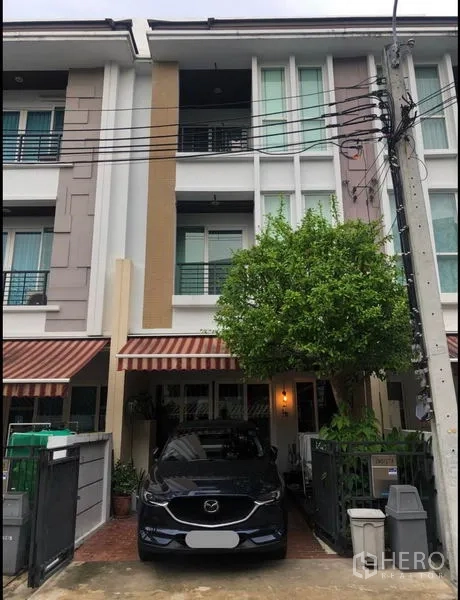 联排别墅 出售在Wang Thonglang, Bangkok - 三层联排别墅外立面，带院内车位、条纹遮阳篷与绿树。