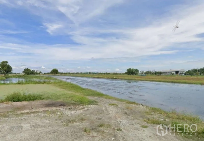 Land for sale in Chachoengsao - Elongated pond with grassy banks and distant low-rise buildings on the horizon.