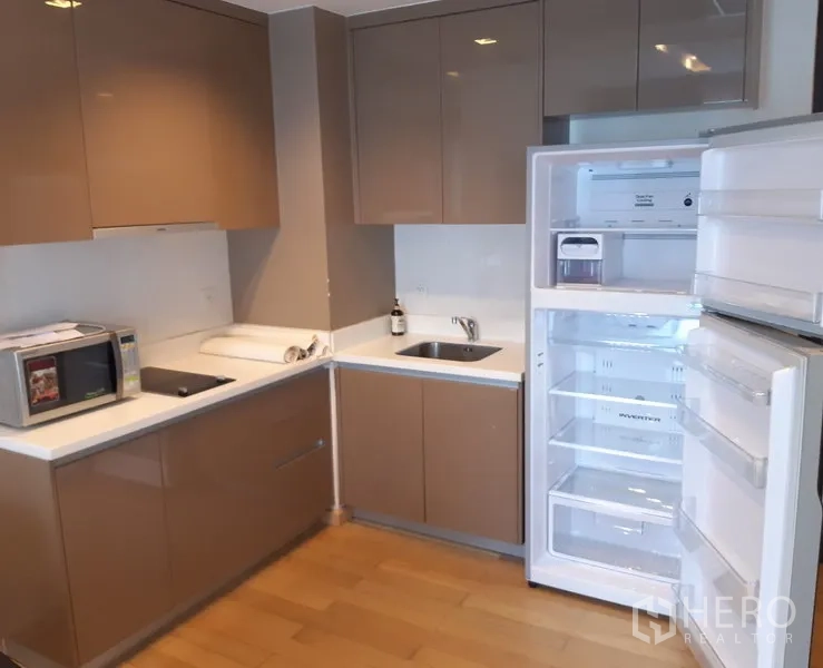 Condominium for rent in Khlong Toei, Bangkok - Modern L-shaped kitchen with glossy cabinets, sink, cooktop, microwave and an open refrigerator.